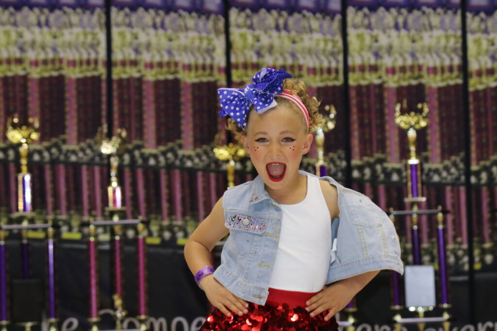 A young girl joyfully posing with a big blue bow and colorful outfit.