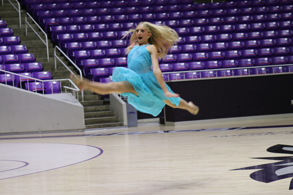 Dancer in a blue dress performs a high leap on a gymnasium floor.