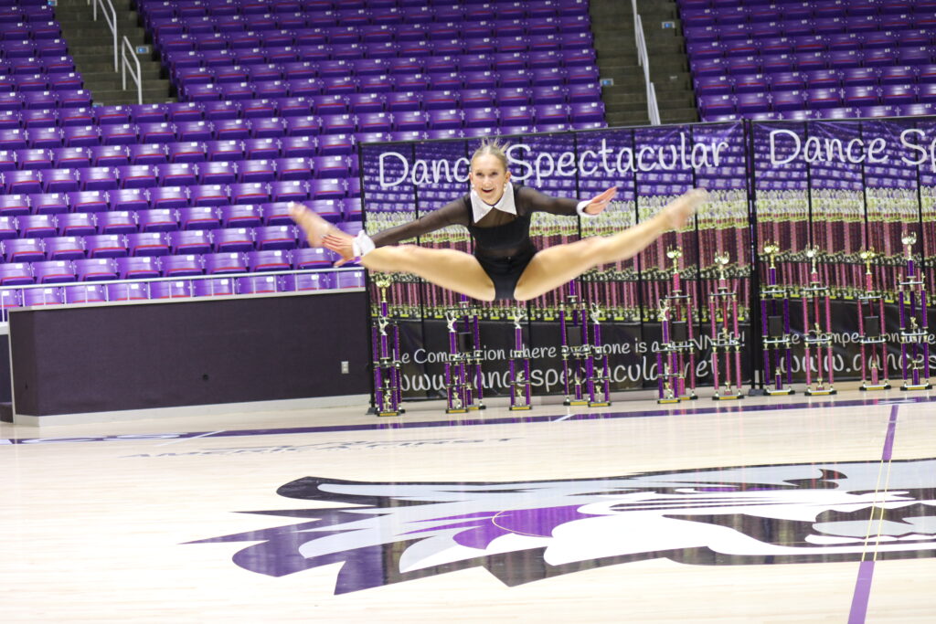 A dancer leaps high in a stadium, wearing a mask and black outfit.